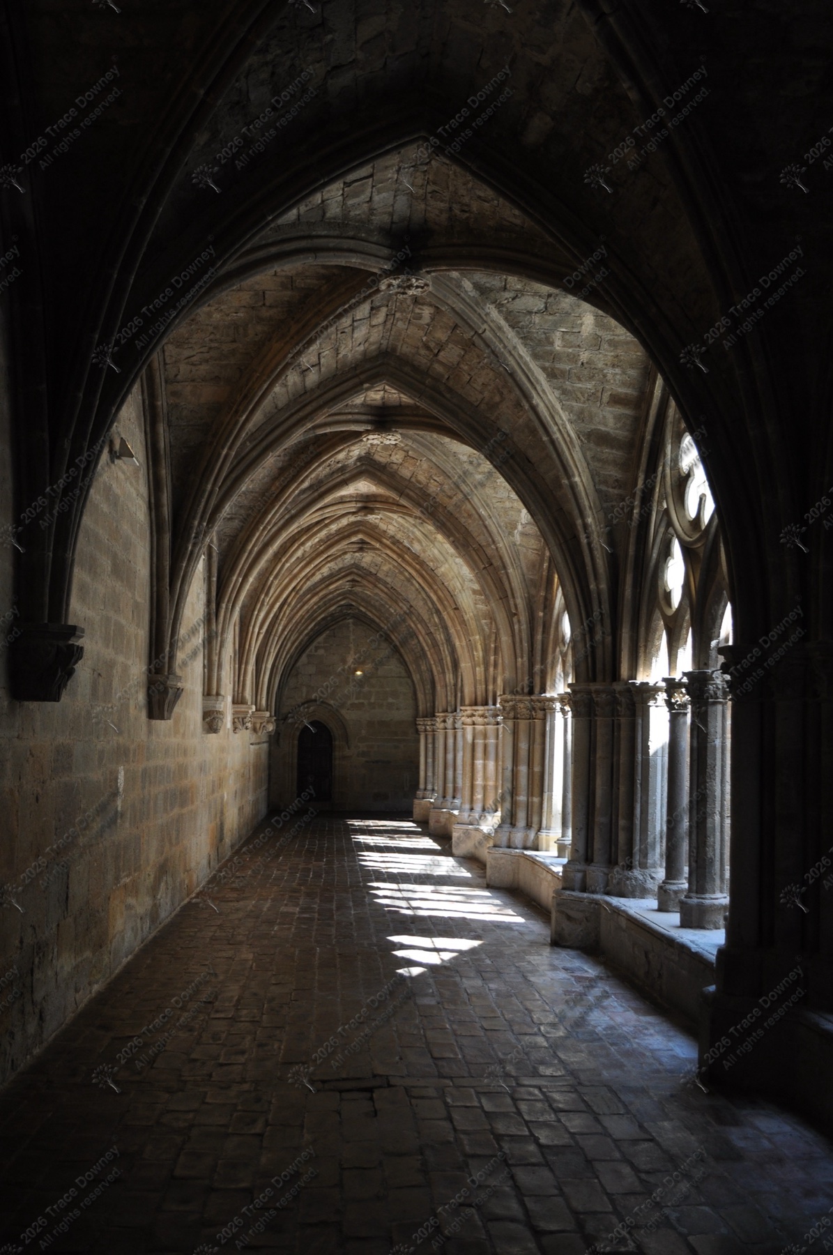Stone cloister corridor with repeating Gothic arches and angled sunlight on the floor.